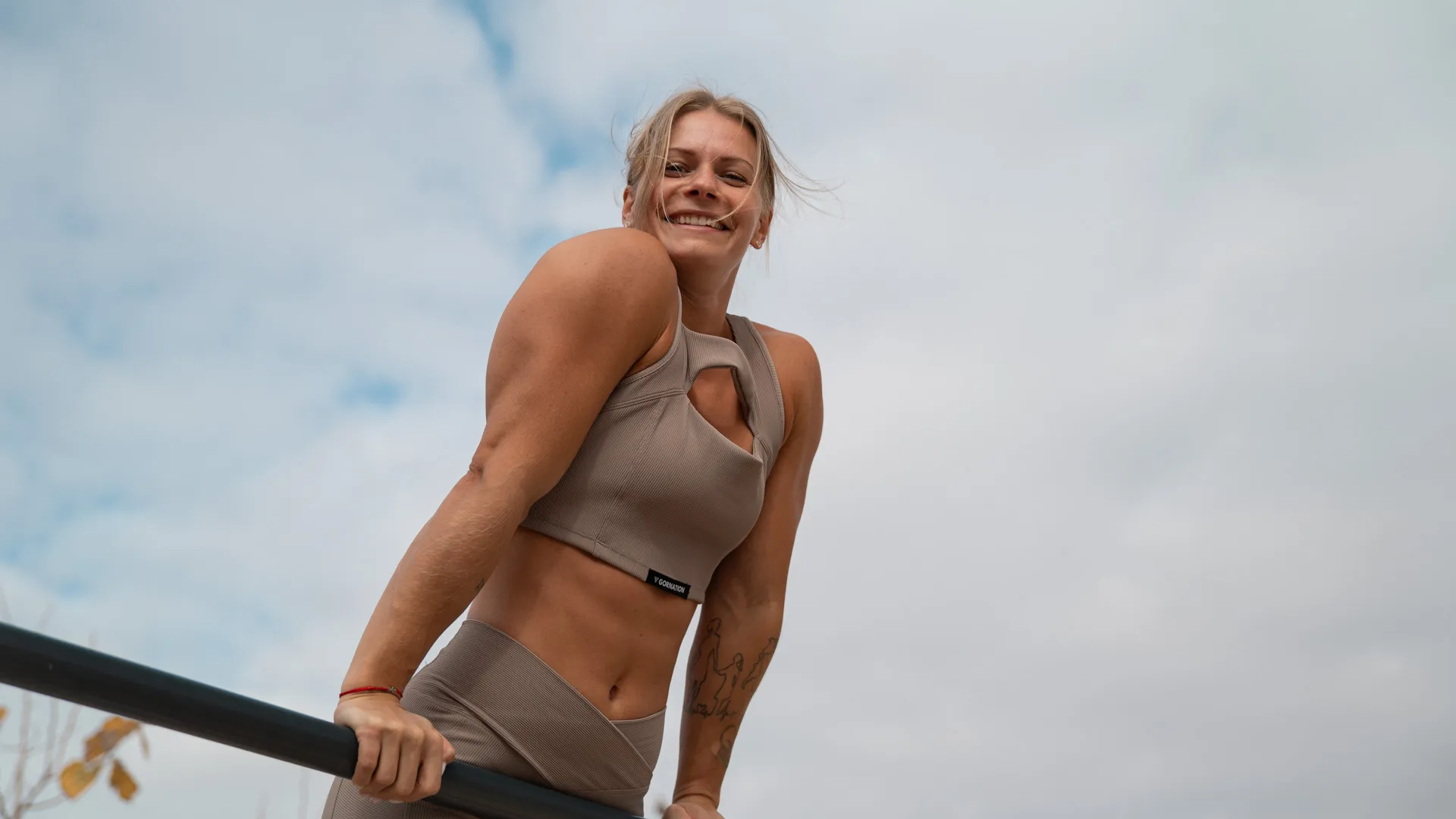 Woman performing a dip exercise on parallel bars, smiling and exuding confidence, while wearing a beige workout set. The background features a cloudy sky, highlighting her strength and dedication to calisthenics training in an outdoor environment.